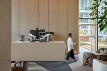 Load image into Gallery viewer, A person walks past a Useabot Dual-Arm Robotic Coffee Station by Elite Robots (Open-Style Workbench) in a bright, minimalist lounge with wooden walls, large windows, and stylish seating.
