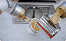Load image into Gallery viewer, The Useabot Dual-Arm Robotic Coffee Station by Elite Robots (Enclosed Type or Open-Style Workbench) delivers a cutting-edge self-service coffee experience, pouring coffee from a metallic jug into a white paper cup over a metal-grate counter.
